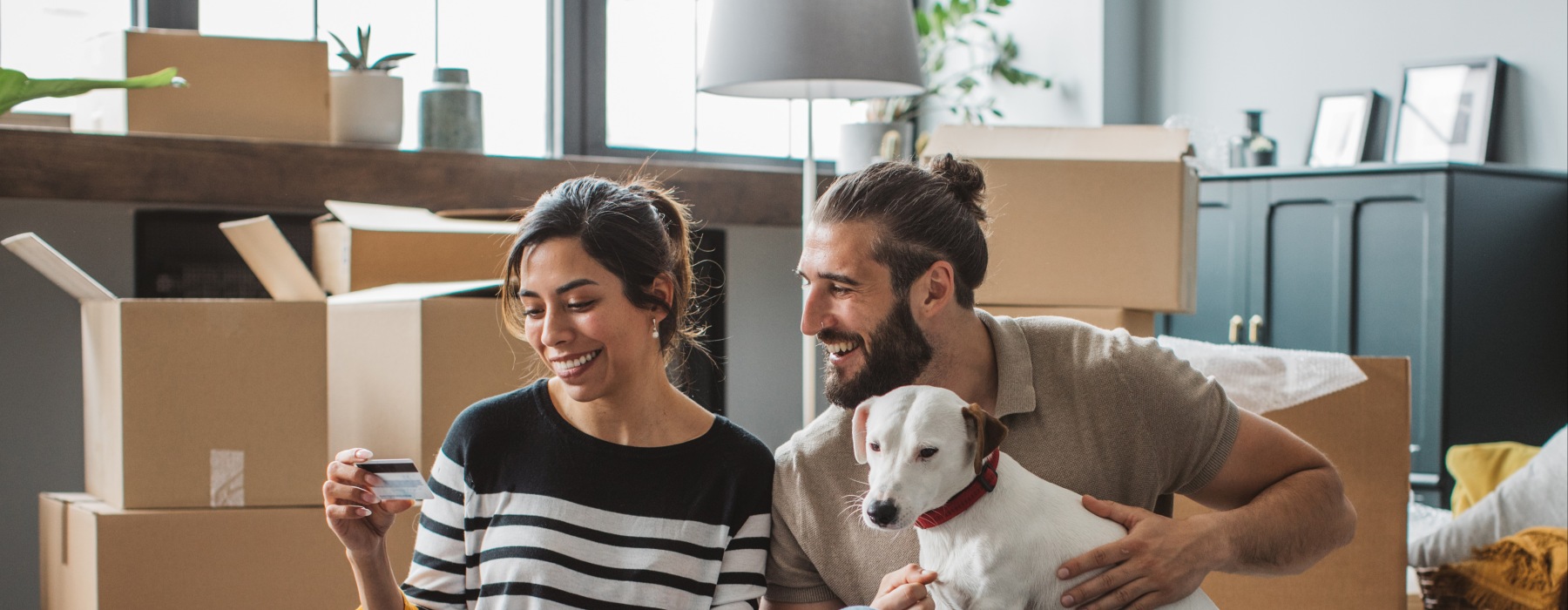 A smiling couple sitting on the floor of their new pet-friendly apartment surrounded by moving boxes, using a laptop and credit card with their dog.